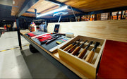 A wooden workbench with an open drawer holding several chisels, shown in a workshop environment with a wood ceiling and industrial shelving in the background.